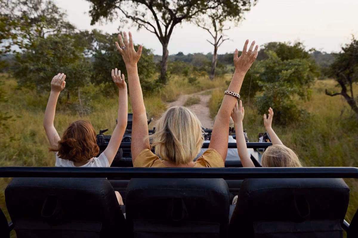 Three people raising hands while seated in an open safari vehicle on a dirt road through trees.