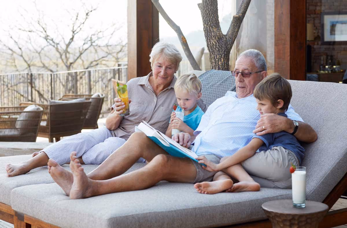 Grandparents sitting on outdoor sofa reading a book with two young boys drinking milk.