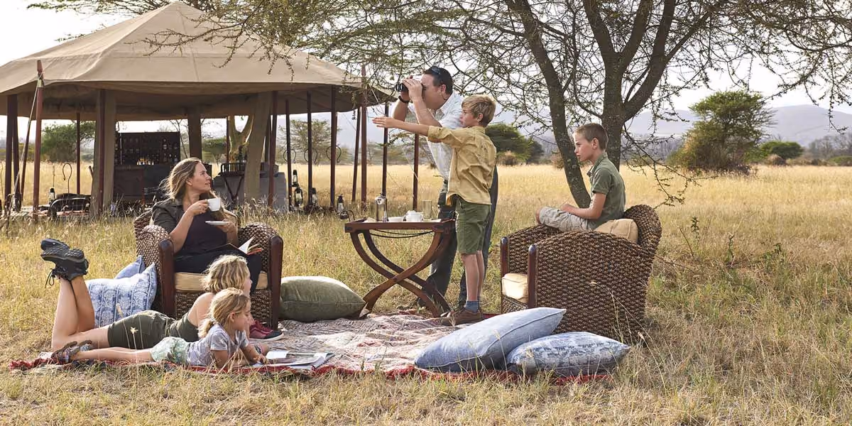 A family enjoying tea and reading outdoors near a tent in a grassy savanna setting.
