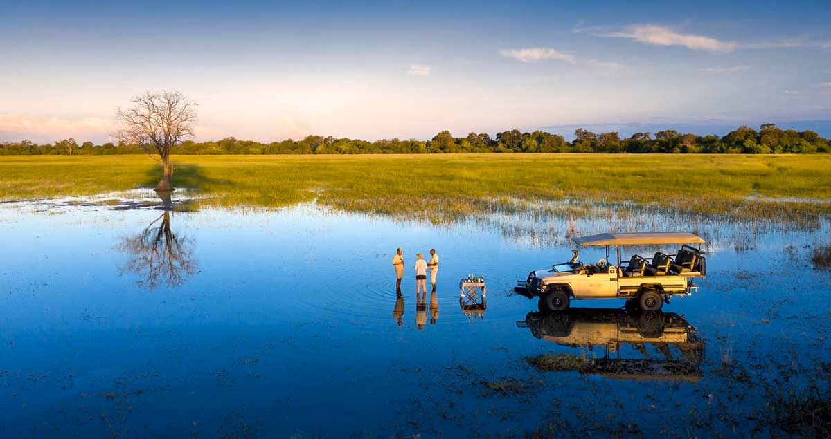 Three people standing in shallow water near a safari vehicle and a lone tree during sunset.