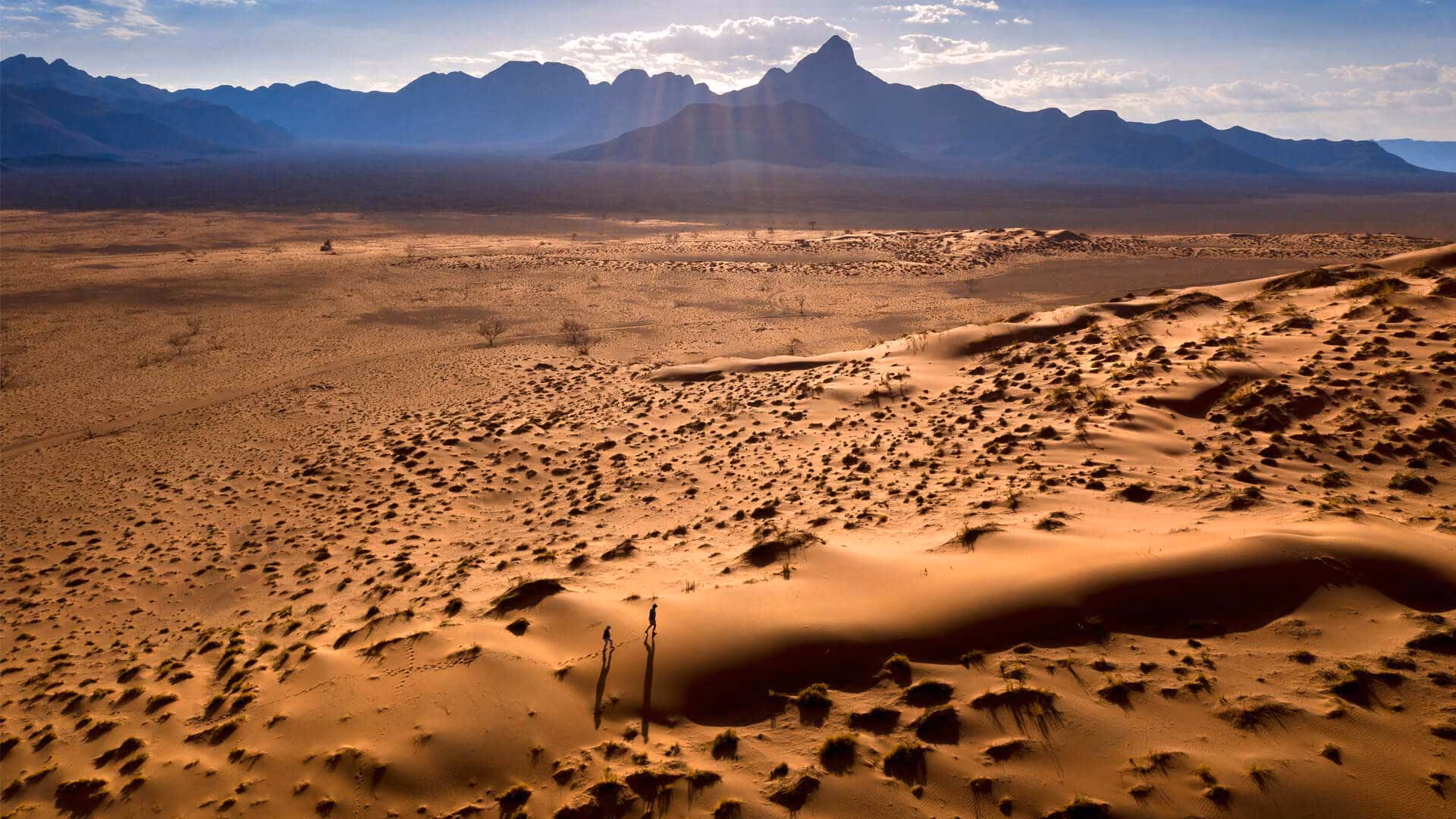 Two people walking on sandy desert dunes with mountains in the distance under a partly cloudy sky.