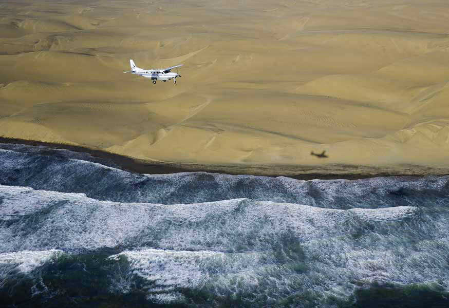 Small white plane flying above sandy beach with waves crashing below and plane's shadow on sand.