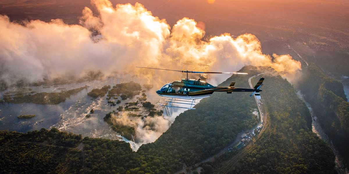 Helicopter flying over a lush green landscape with mist and waterfall under a warm sunset sky.