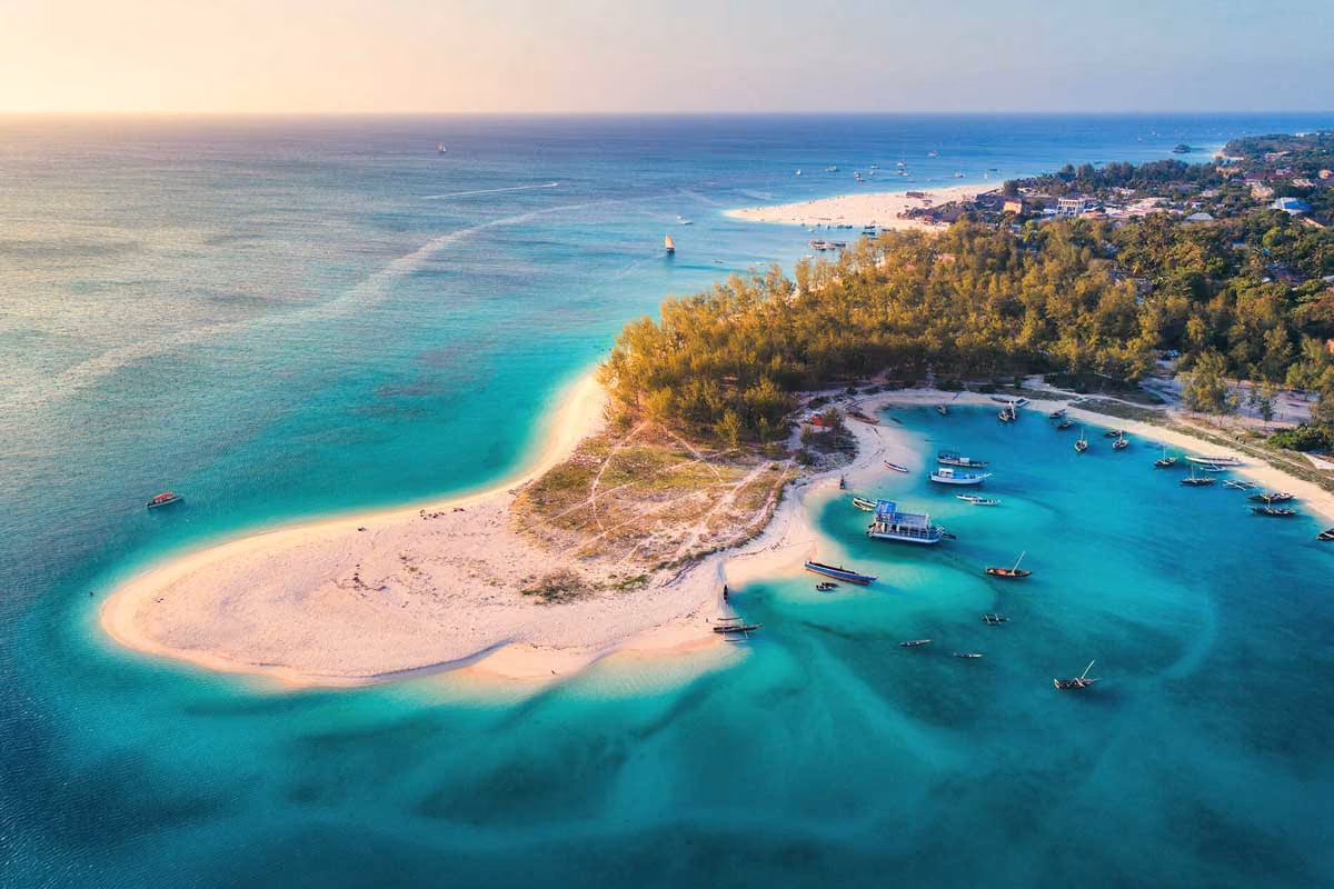 Aerial view of a sandy island with turquoise waters, boats, and dense trees near a coastal town.