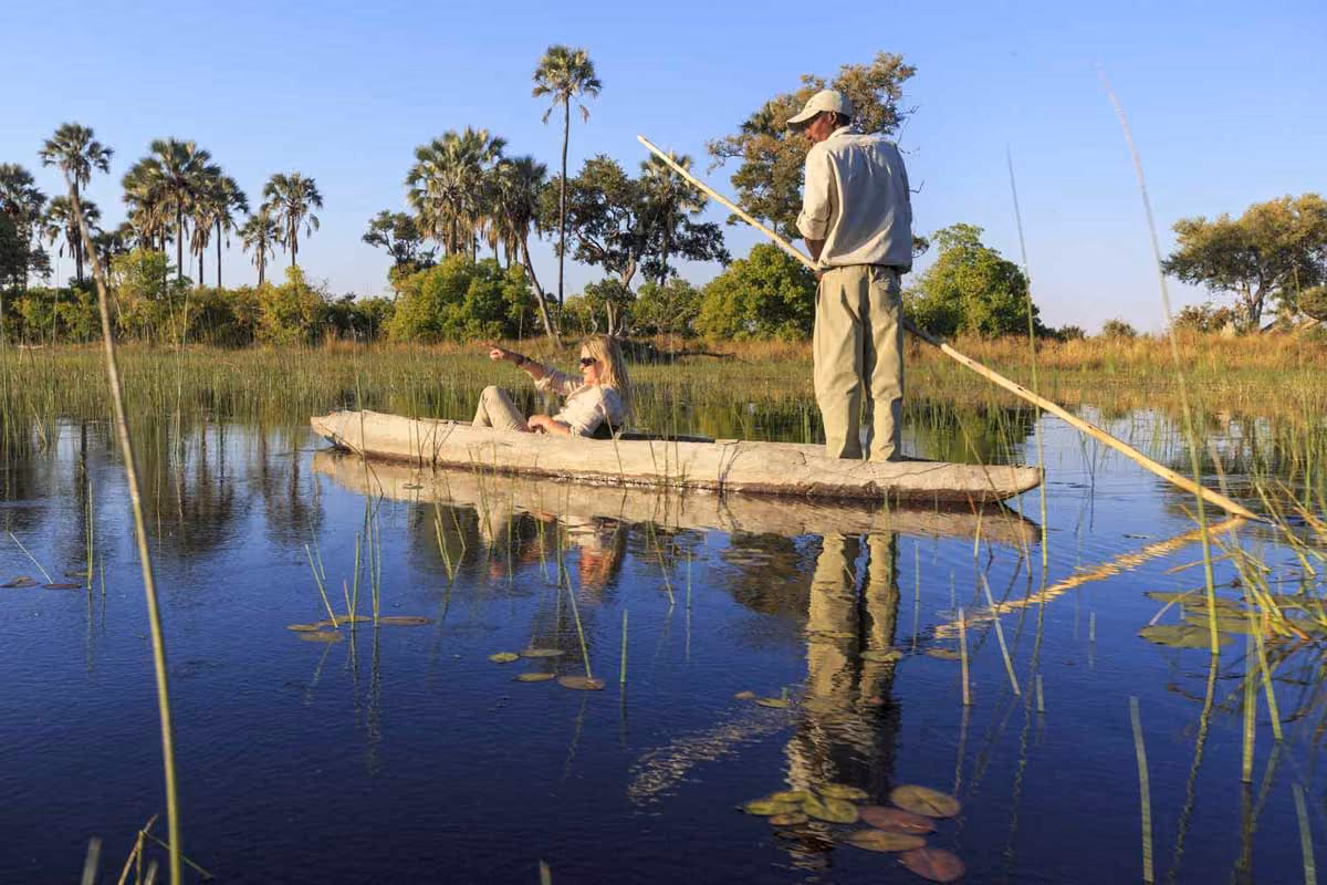 Two people on a wooden canoe on calm water with reeds and palm trees in the background.