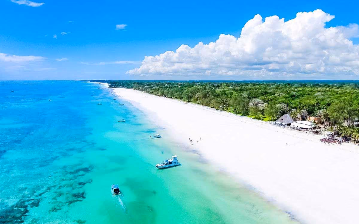 Aerial view of a white sandy beach with turquoise water, boats, and green trees under blue sky.