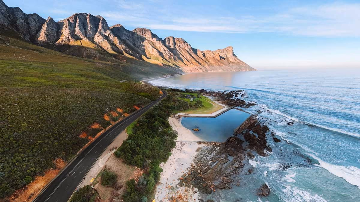 Coastal road alongside rocky mountains with a tidal pool and beach by the ocean under a blue sky.