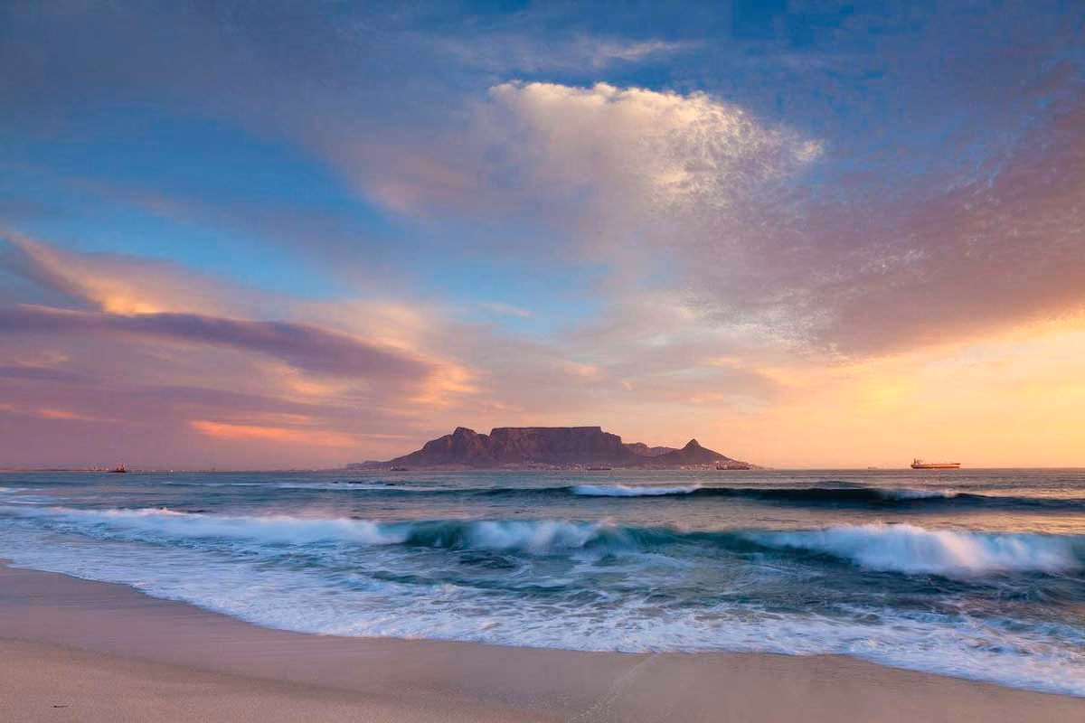 View of a sandy beach and ocean waves with a flat-topped mountain under a colorful sunset sky.