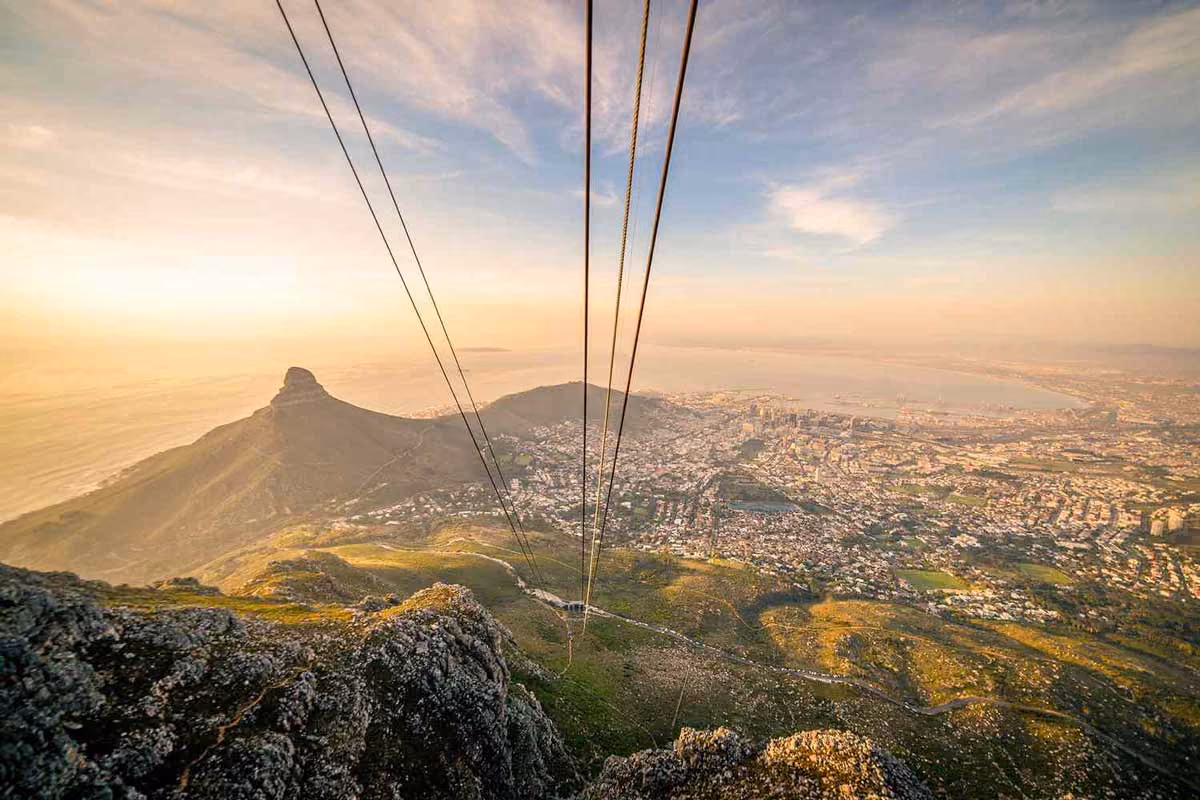 Cable car lines descending a rocky mountain overlooking a city and coastline at golden hour.