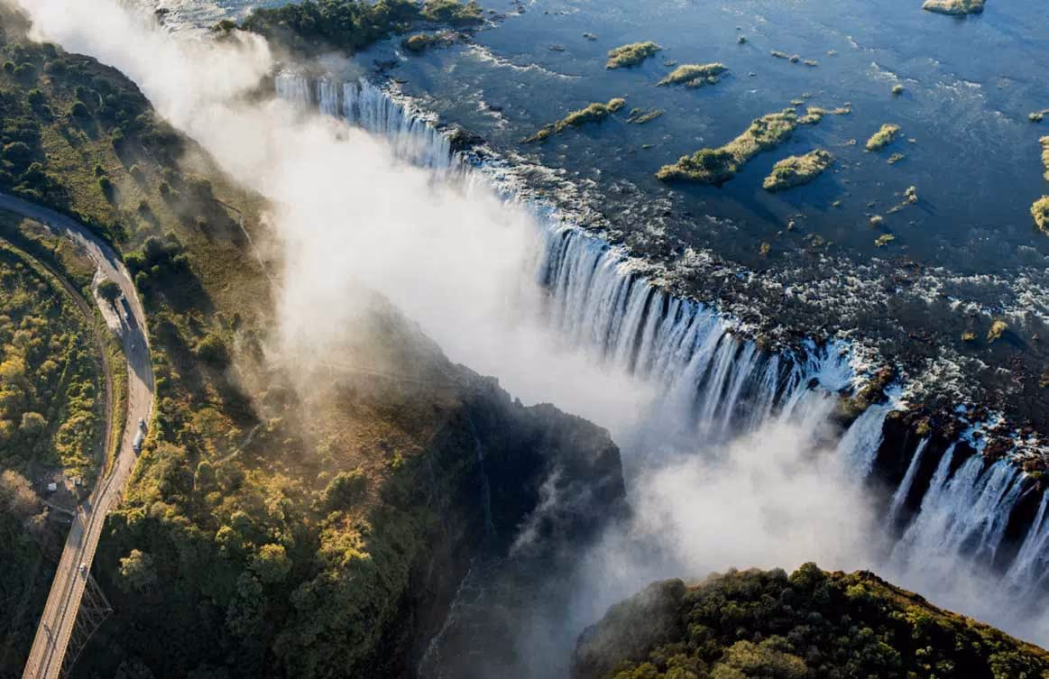 Aerial view of a large waterfall with mist rising and a winding road nearby through green terrain.