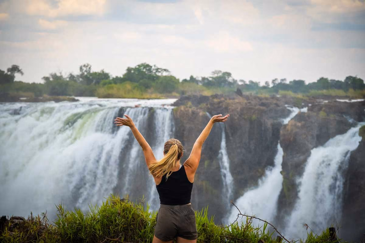 Woman with arms raised facing a large waterfall surrounded by greenery and rocky cliffs.