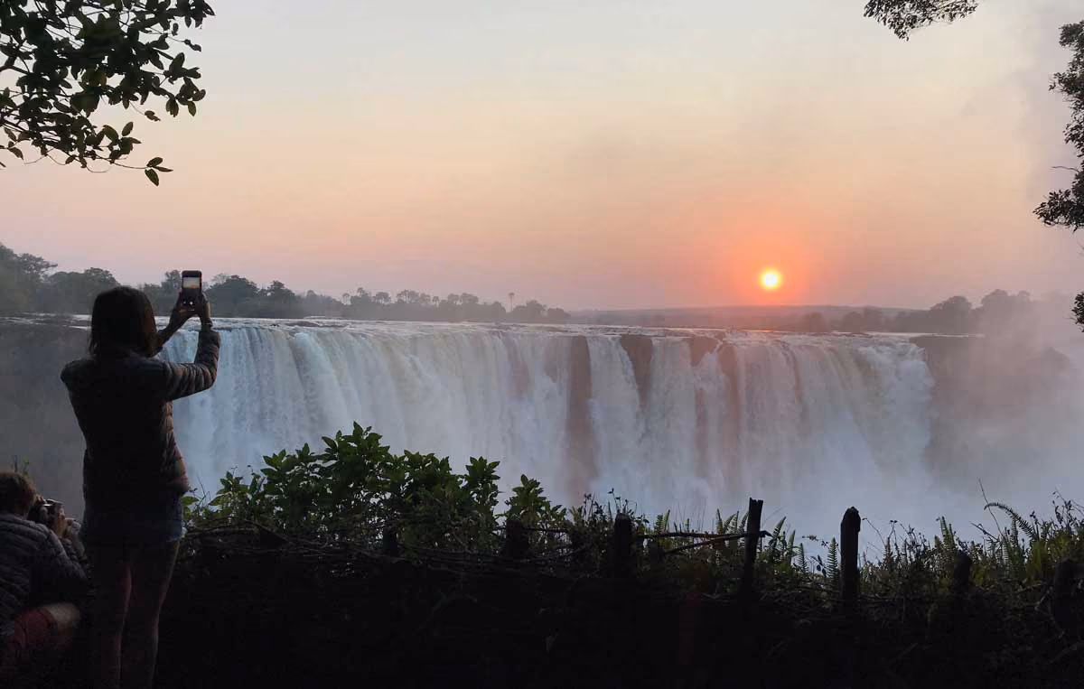 Silhouettes of two people capturing photos of a wide waterfall at sunset with mist and trees around.
