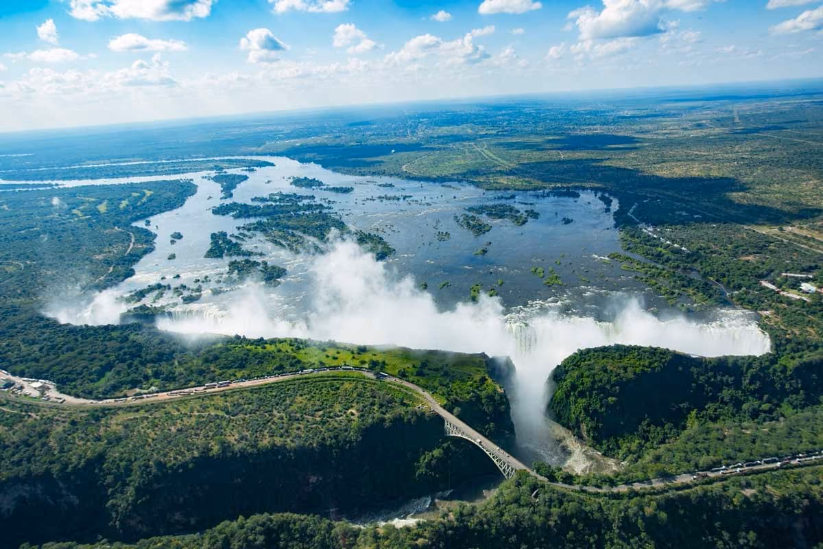 Aerial view of a large waterfall with mist and a bridge crossing a gorge surrounded by lush greenery.