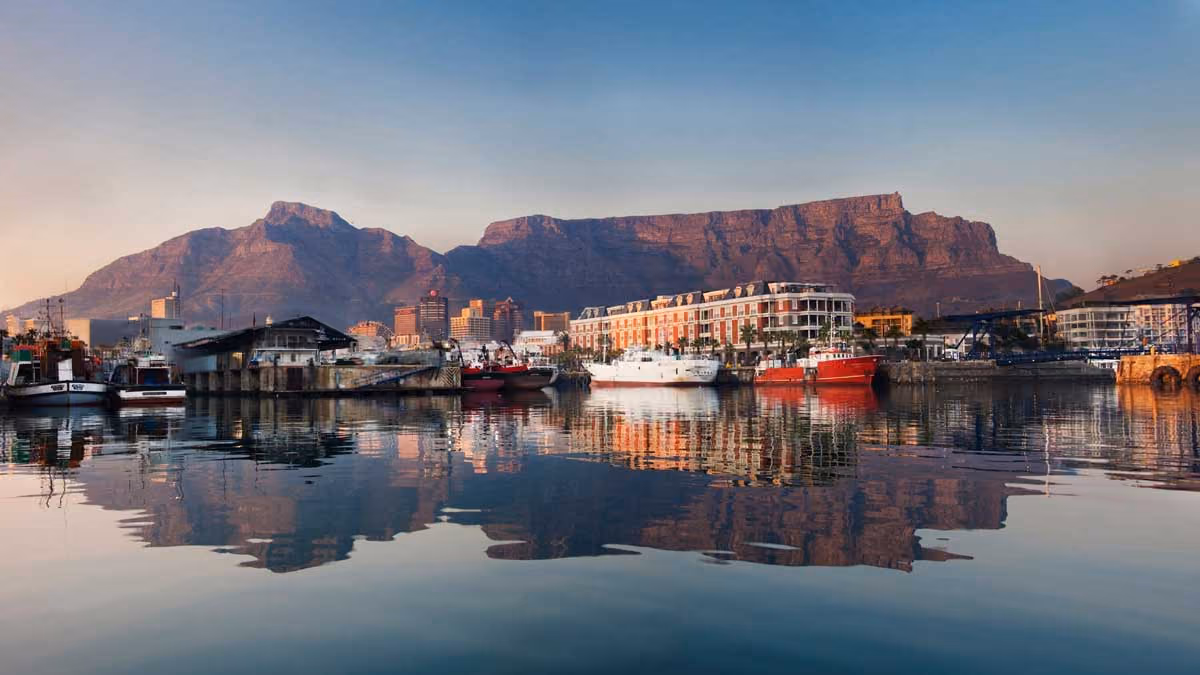 Harbor with boats and waterfront buildings reflected in calm water, with large flat-topped mountain in background.