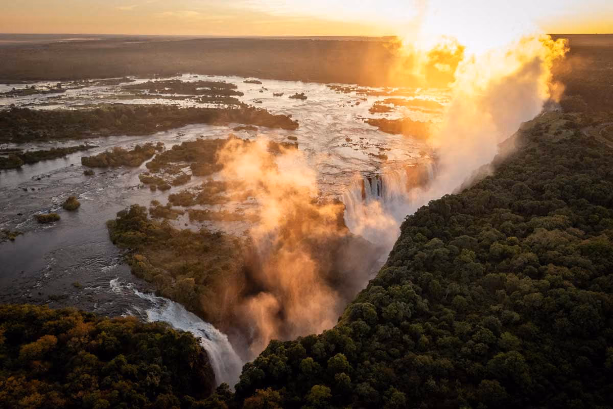 Sunset over a large waterfall with mist rising and lush green forest on the sides.