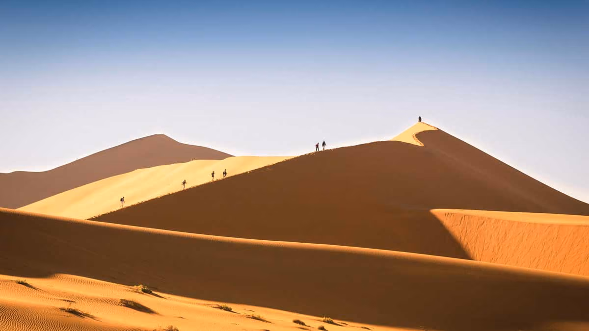 People walking along the ridge of tall sand dunes under a clear blue sky in a desert.