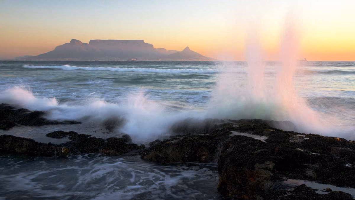 Waves crashing against rocky shore with a flat-topped mountain and sunset in the background.