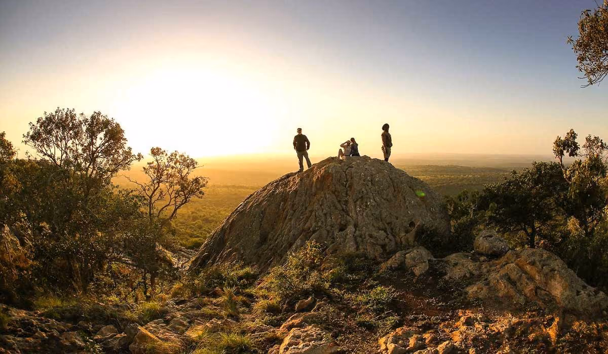 Three people standing and sitting atop a rocky hill at sunset with trees and forest below.