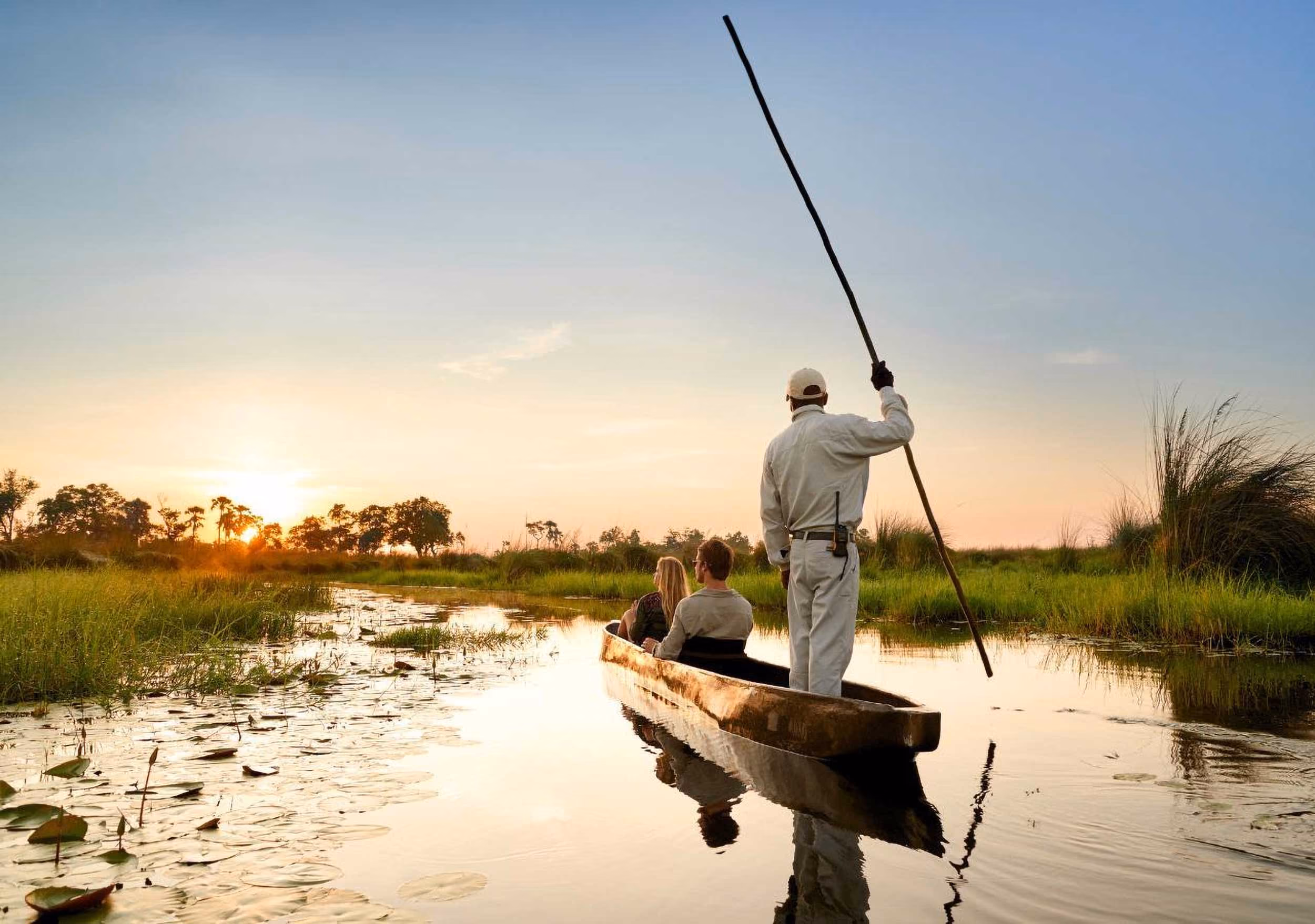 Three people in a wooden canoe on calm water surrounded by grass at sunset.
