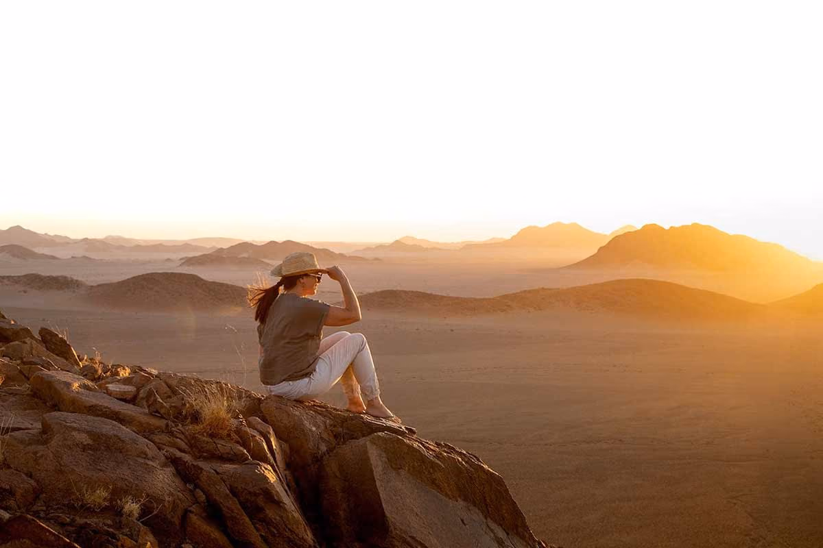 Woman wearing hat sitting on rocky hill watching sunlit desert landscape with mountains at sunset.