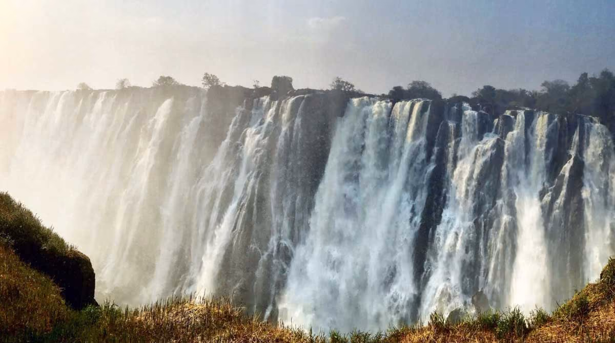Large waterfall cascading over a rocky cliff with mist rising and vegetation in the foreground.