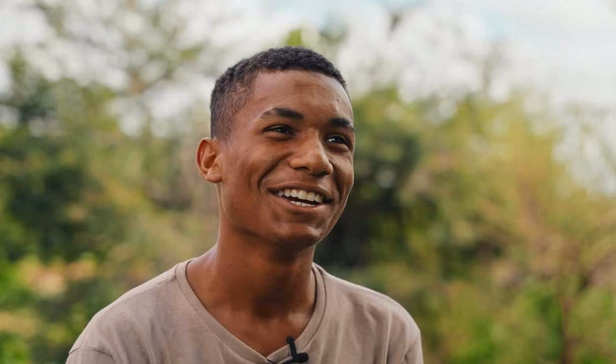 Smiling young man wearing a beige shirt in an outdoor setting with blurred greenery background.