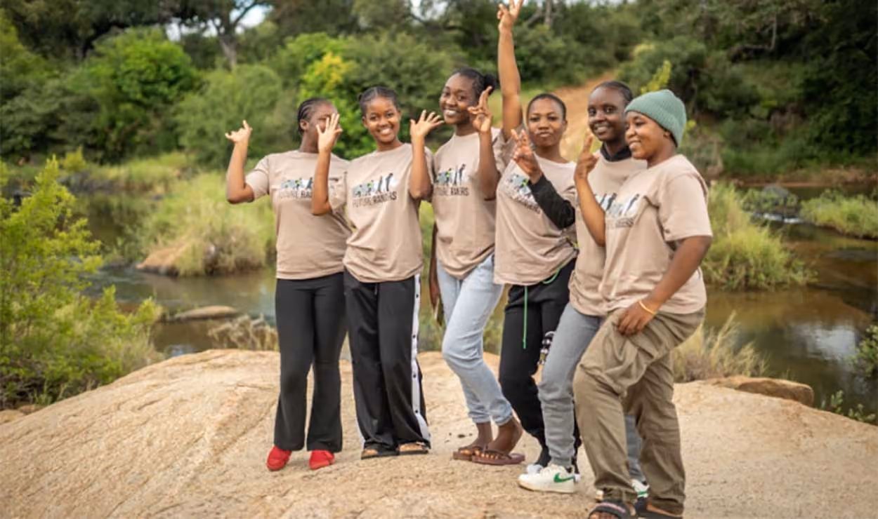 Six young women standing on a rock near water, waving and smiling, wearing matching tan T-shirts.