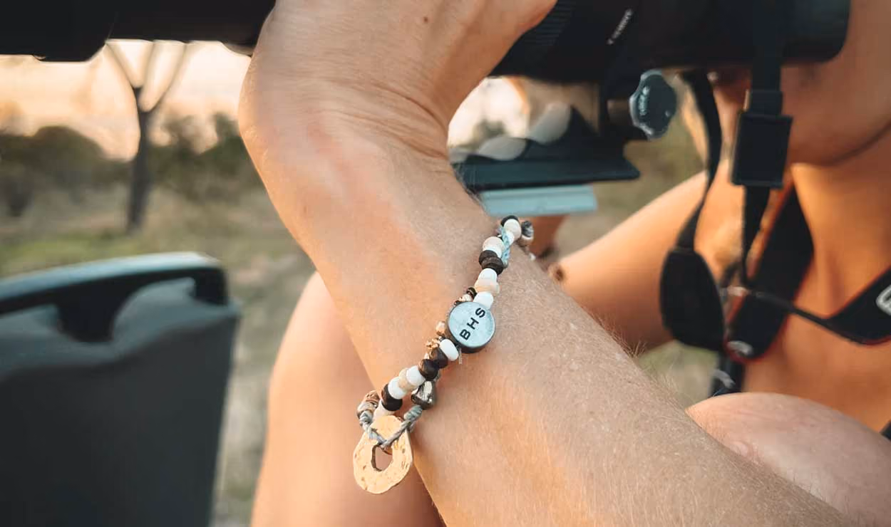 Person wearing a beaded bracelet with a BHS charm while looking through binoculars outdoors.
