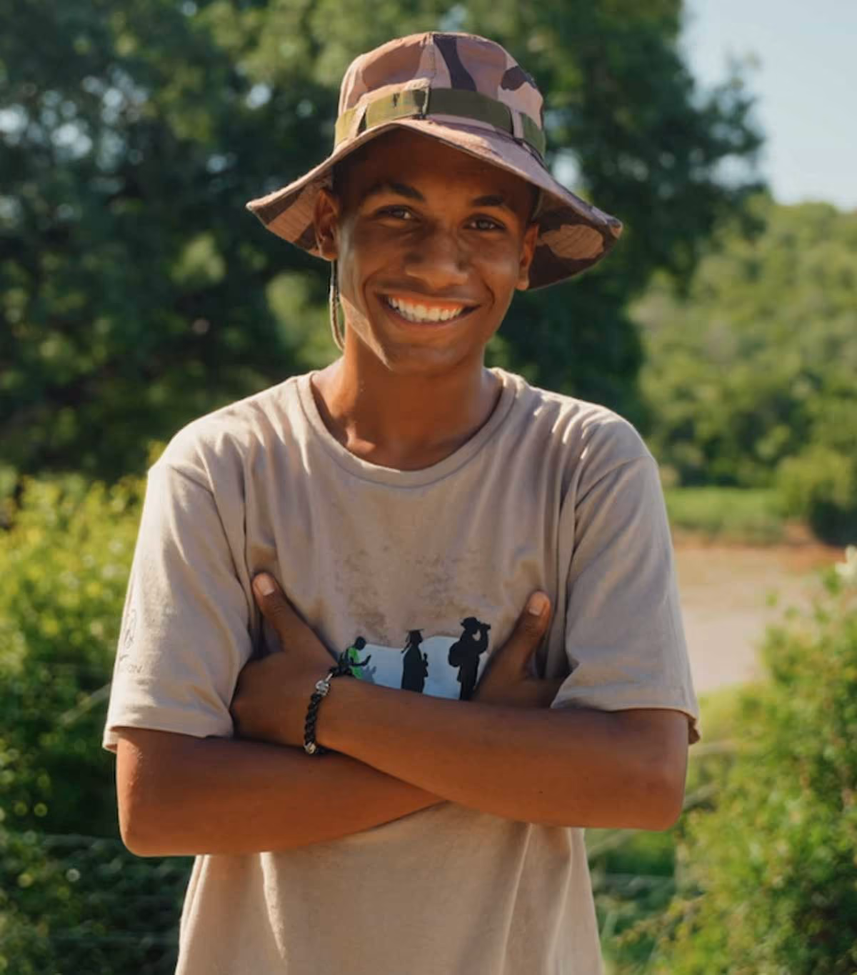 Smiling young man wearing a camouflage bucket hat and beige t-shirt standing outdoors with arms crossed.