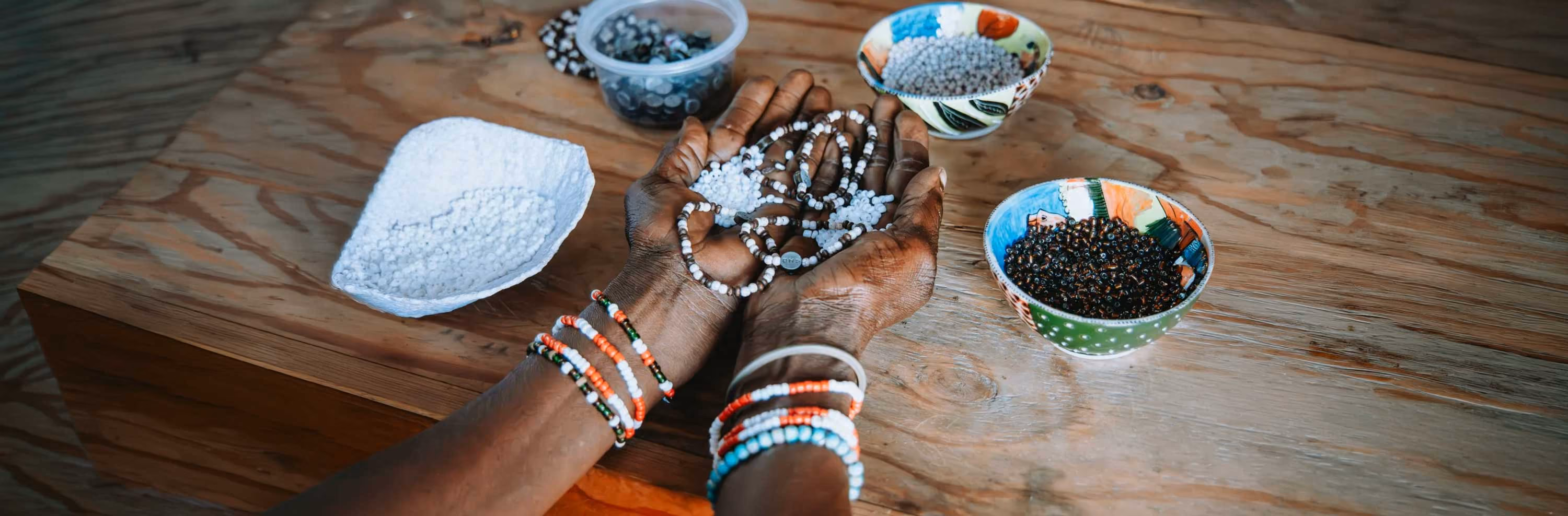 Hands holding white beaded necklaces with bowls of beads on a wooden table, wearing colorful bracelets.