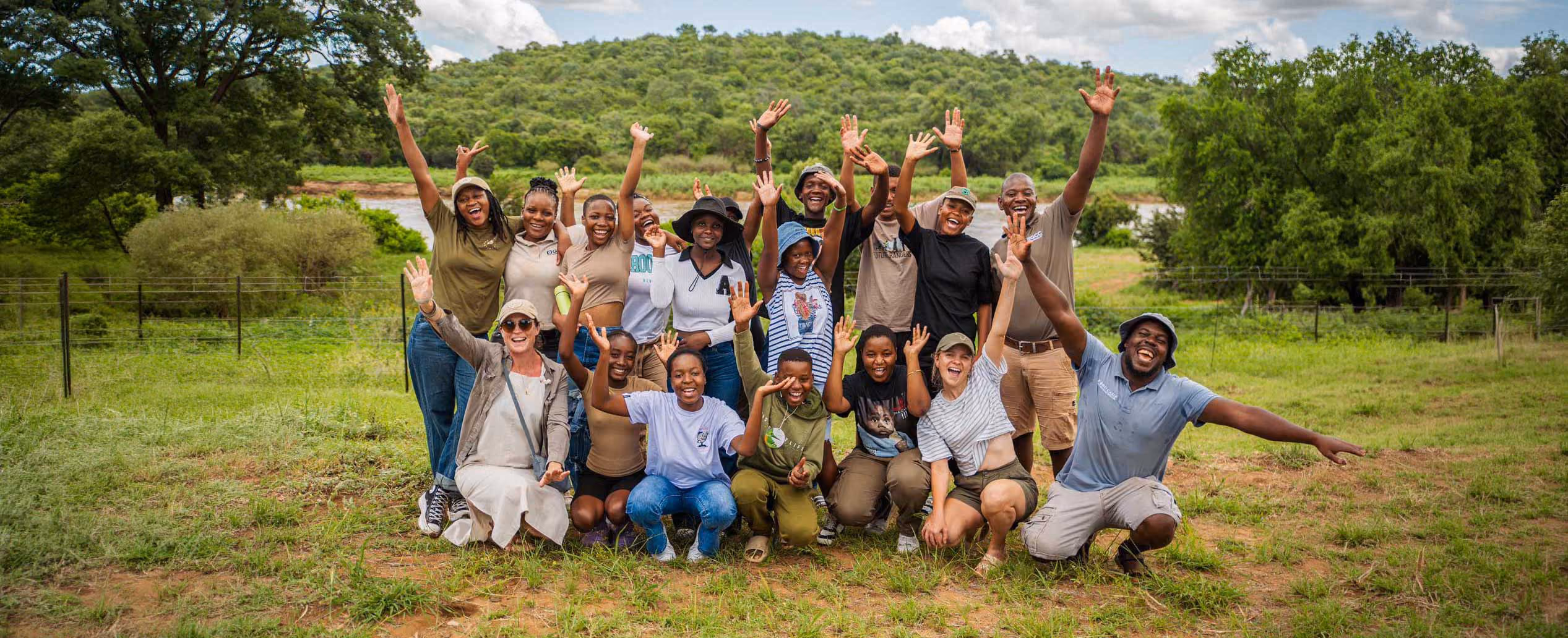 Group of smiling people outdoors in a green field, raising hands happily with trees and hills behind them.