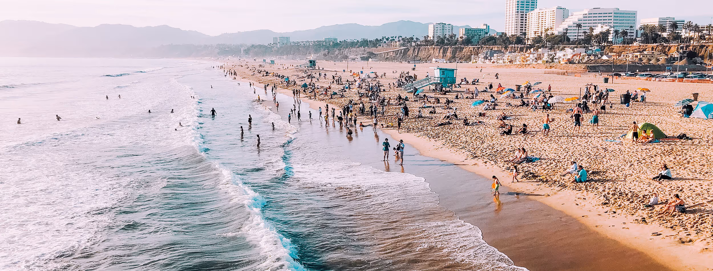 Crowded sandy beach with people sunbathing, playing, and swimming near lifeguard towers and coastal buildings under a hazy sky.
