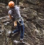 Person wearing an orange helmet and harness rock climbing on a steep rocky surface.