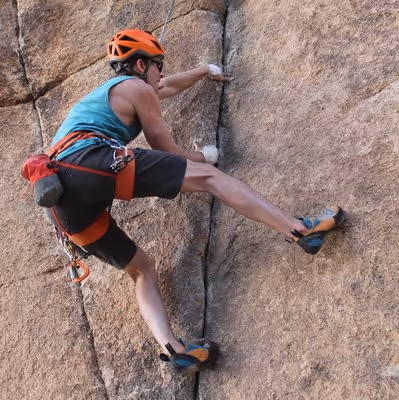 Man wearing an orange helmet and climbing shoes, scaling a large rock face using cracks for hand and footholds.