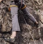 Person climbing a rocky cliff wearing gray pants and climbing shoes with a safety harness and rope.