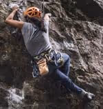 Man wearing an orange helmet climbing a rocky cliff with climbing gear and chalk bag.