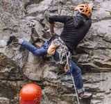 Climber wearing an orange helmet and black shirt scaling a rocky cliff with climbing gear attached.