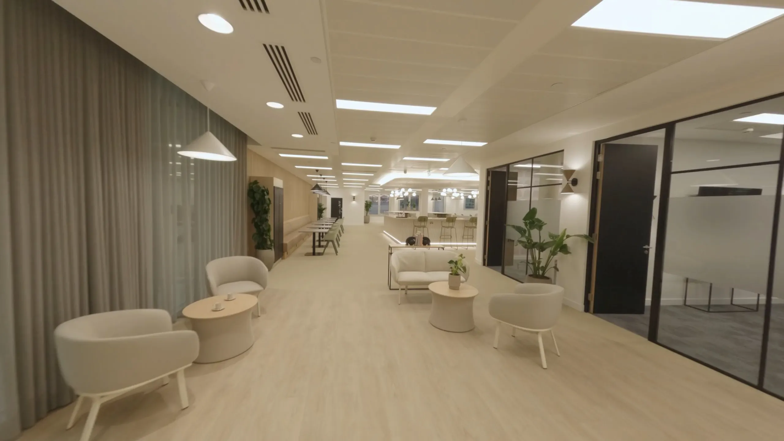 Modern, spacious office lounge with beige chairs, small round tables, plants, and glass-walled meeting rooms.