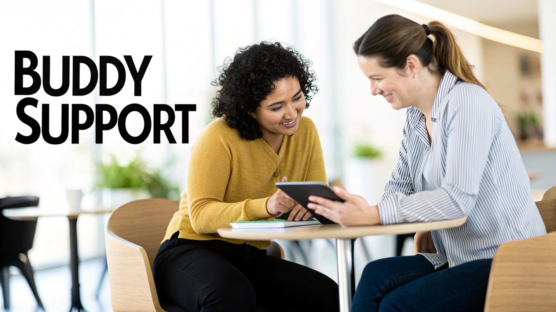 Two smiling women collaborate at a table, one showing content on a tablet to the other.