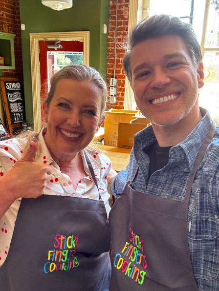 A blond smiling woman gives the thumbs up sign while standing next to a smiling man, both wearing chef aprons that read, “Sticky Fingers Cooking.”