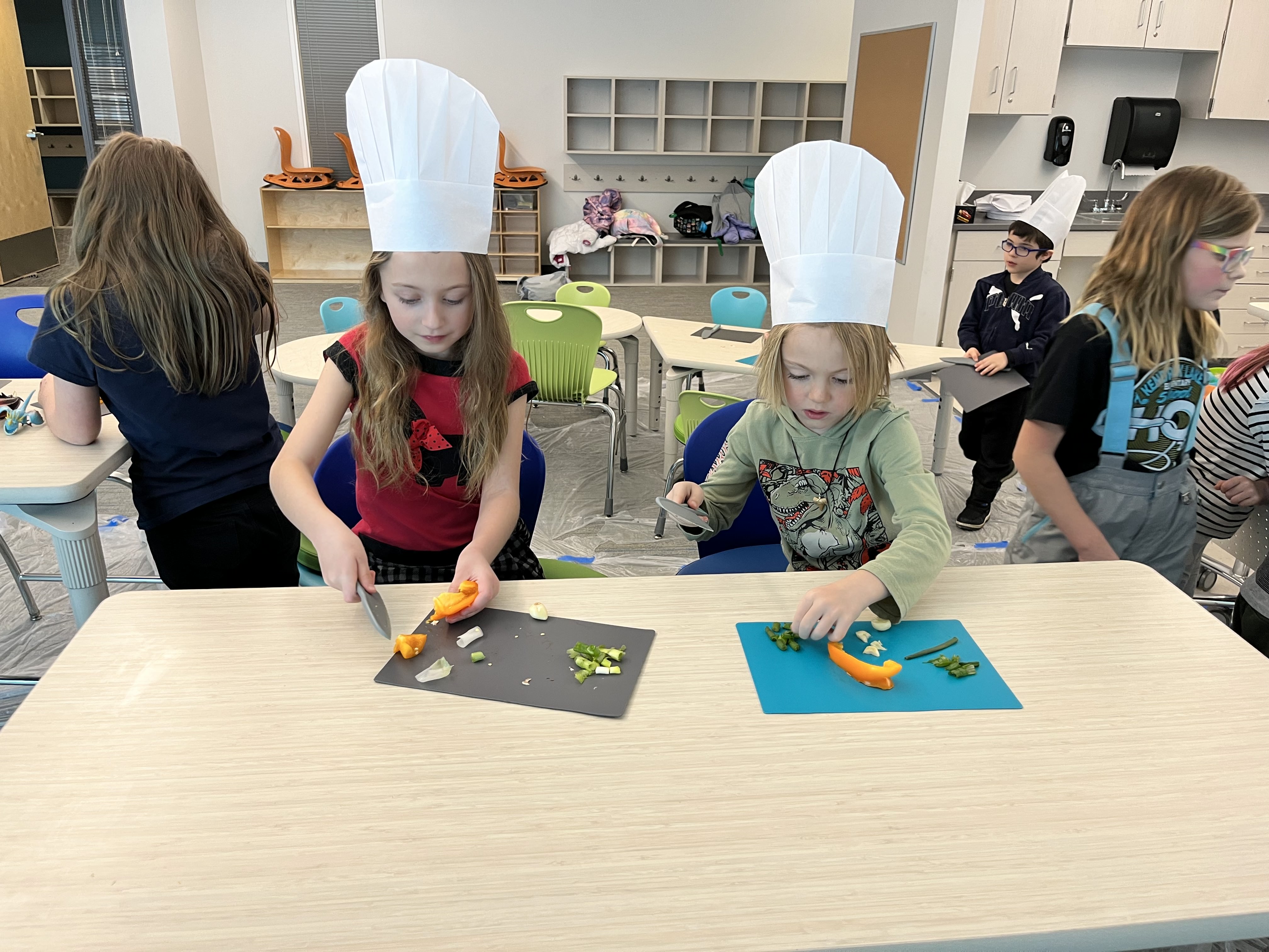 Two girls in a Sticky Fingers Cooking class with chefs hats on are chopping veggies on cutting boards