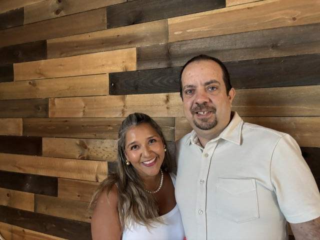 New franchise owners, Sergio and Priscilla, smiling for a photo together in front of a wooden backdrop. Both are dressed in nice clothing