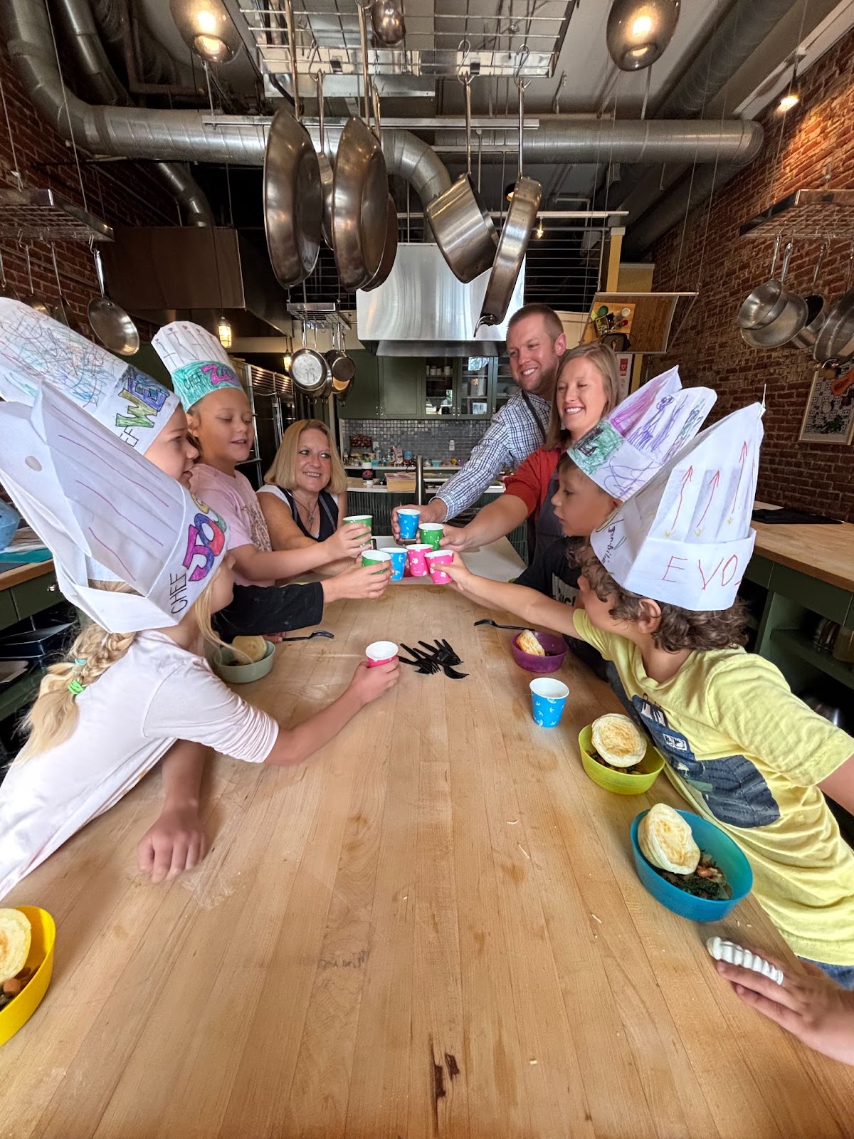Five children wearing white chef hats and three adults make a toast with their colorful paper cups over a wooden countertop.