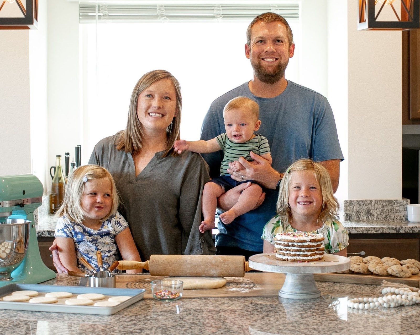 A mom and dad along with three young children smile over a countertop filled with baked goods.