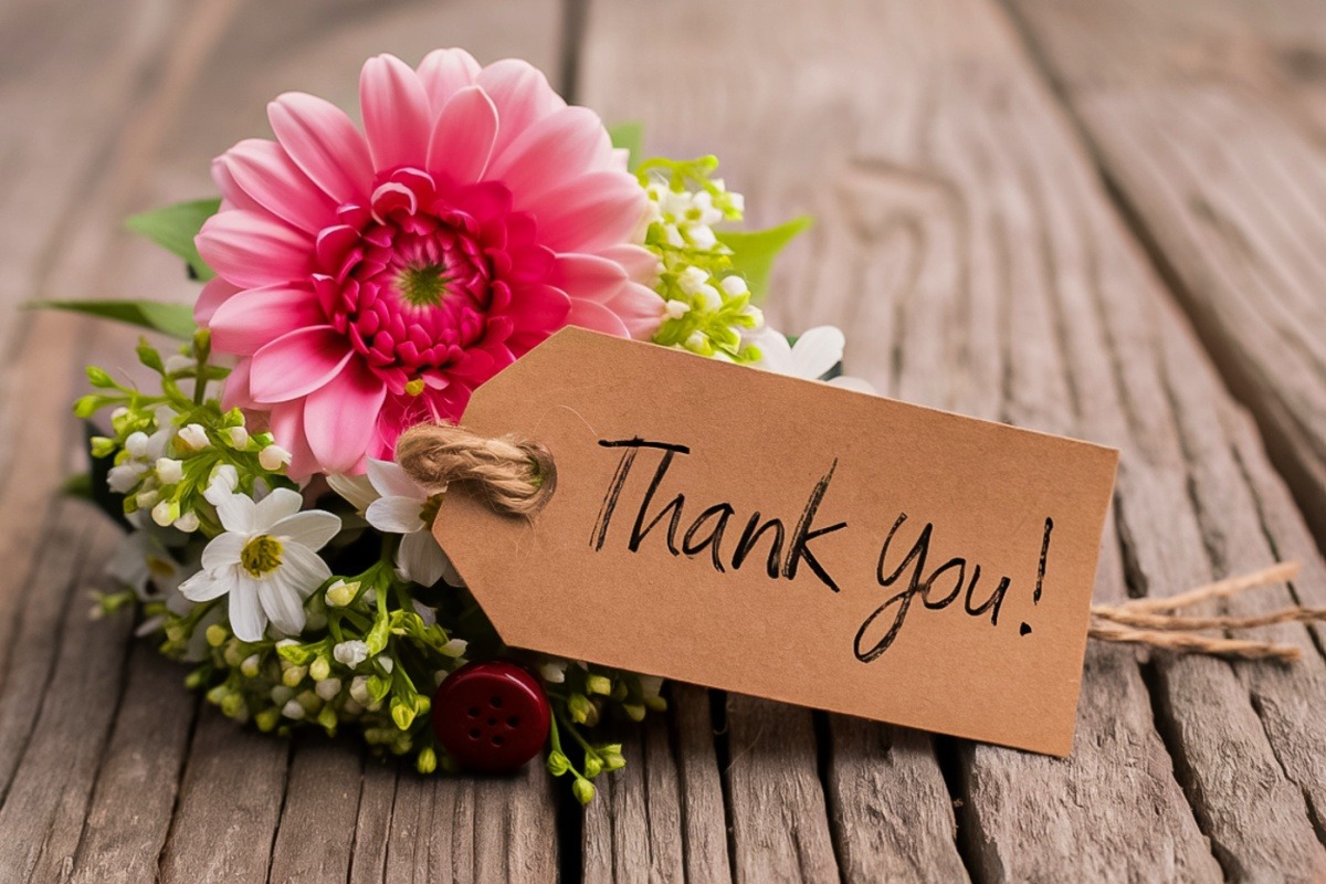 A bouquet of small white flowers and one pink flower sits on a wooden table with a small cards that reads, “Thank you!”