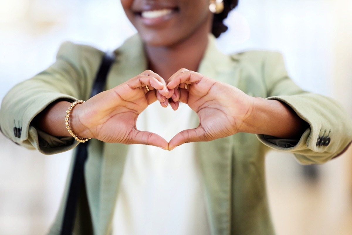 A woman with a gold bracelet and wearing a pale green blazer holds her hands out in front of her to make a heart.