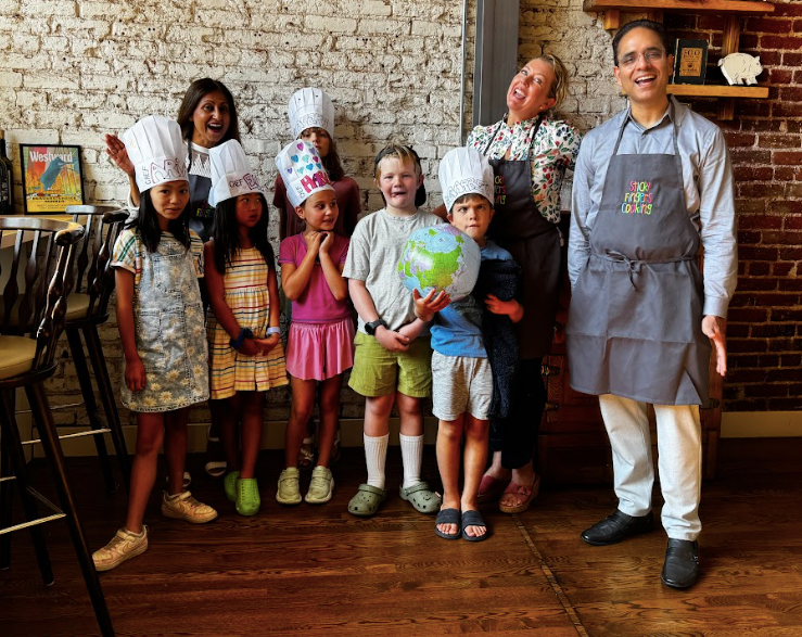 Six young children in white paper chef hats decorated with their names make silly faces for the camera with two smiling women and one smiling man.