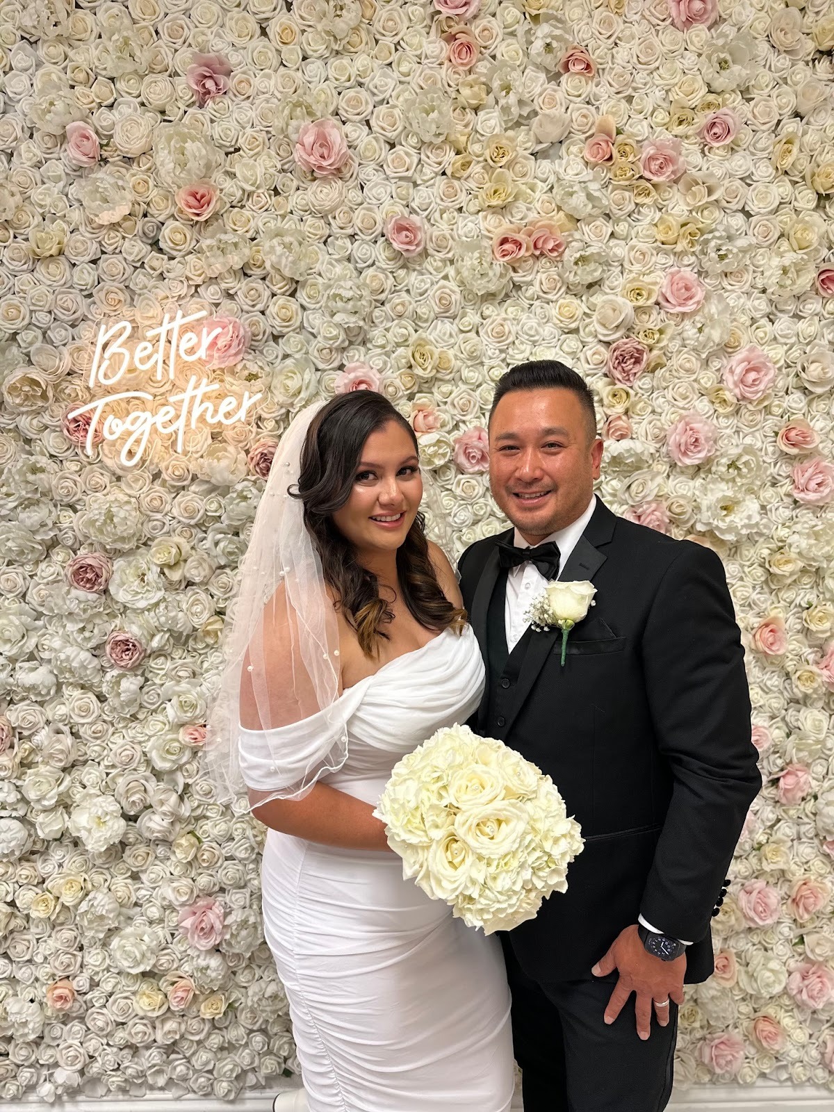 A bride and groom stand smiling in front of a wall of white and pale pink roses with a neon sign that reads, “Better Together.”