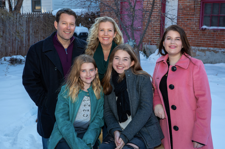 A man and woman with three daughters smile at the camera while standing in the snow. 