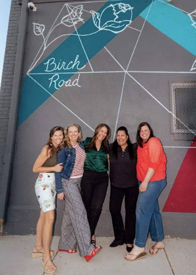 Five women laughing and smiling in front of a gray, blue, and pink wall that reads Birch Road above them.